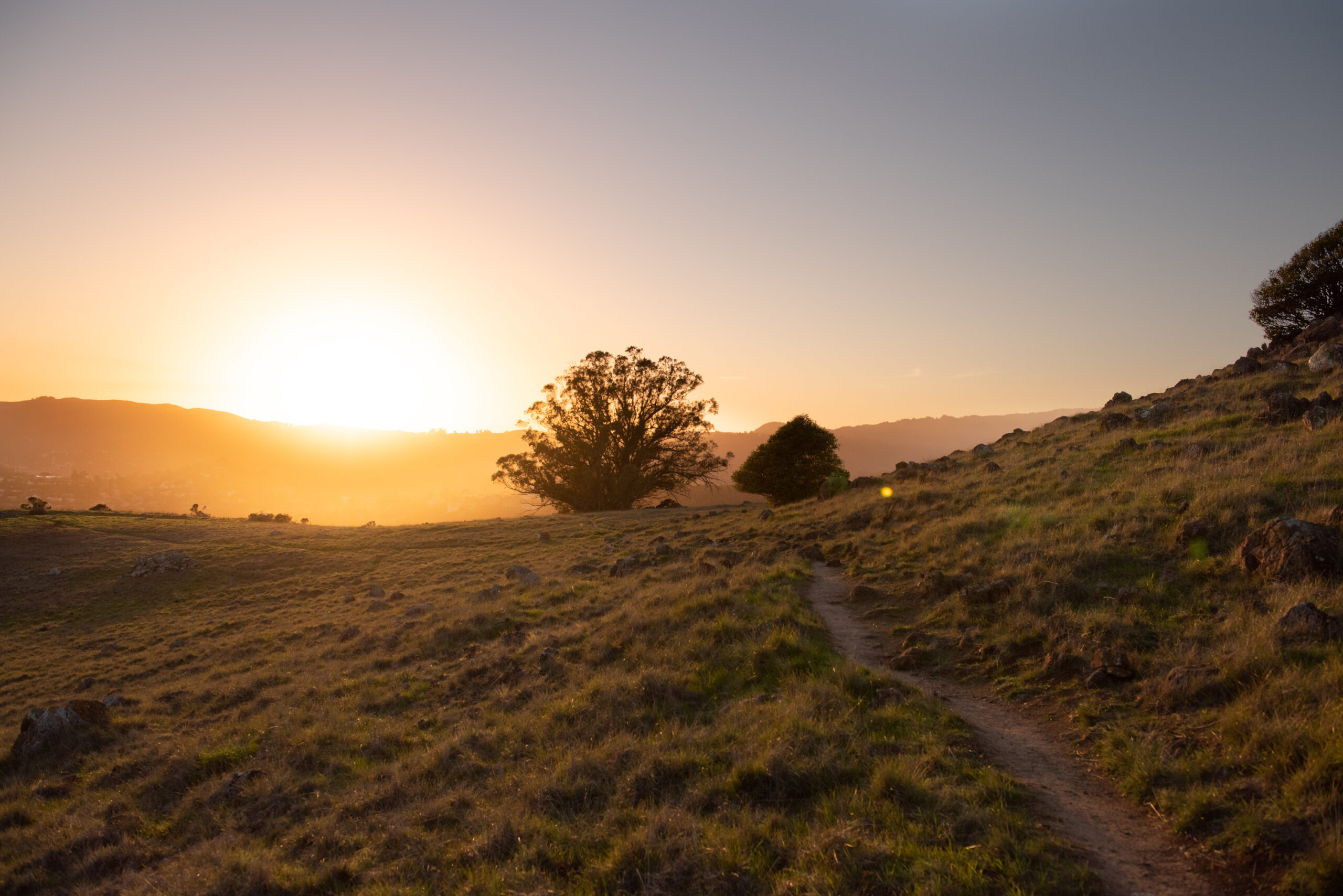 Photo of a trail going along the side of a hill, with more hills and the setting sun in the background