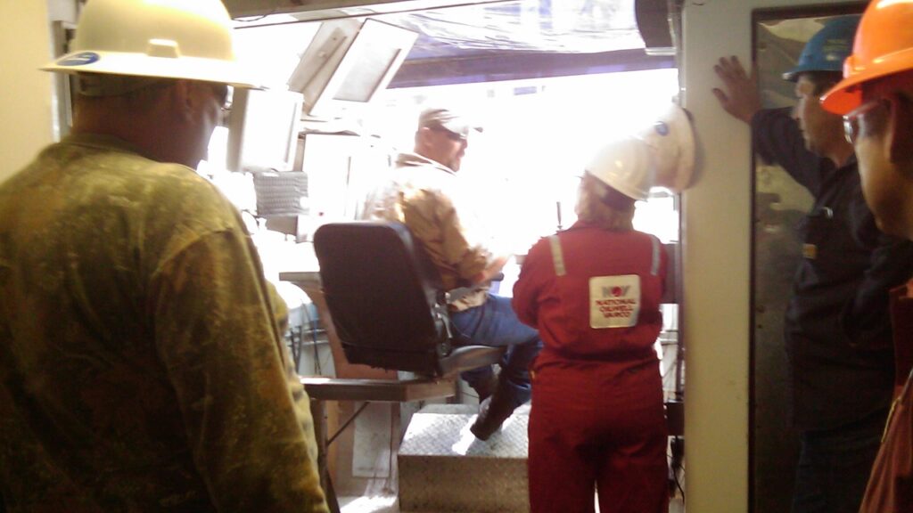 photo of listener (in red coveralls and hardhat) with one of the oilwell operators, seated on the rig, with three screens to his left. Three other people in hard-hats and coveralls are watching.